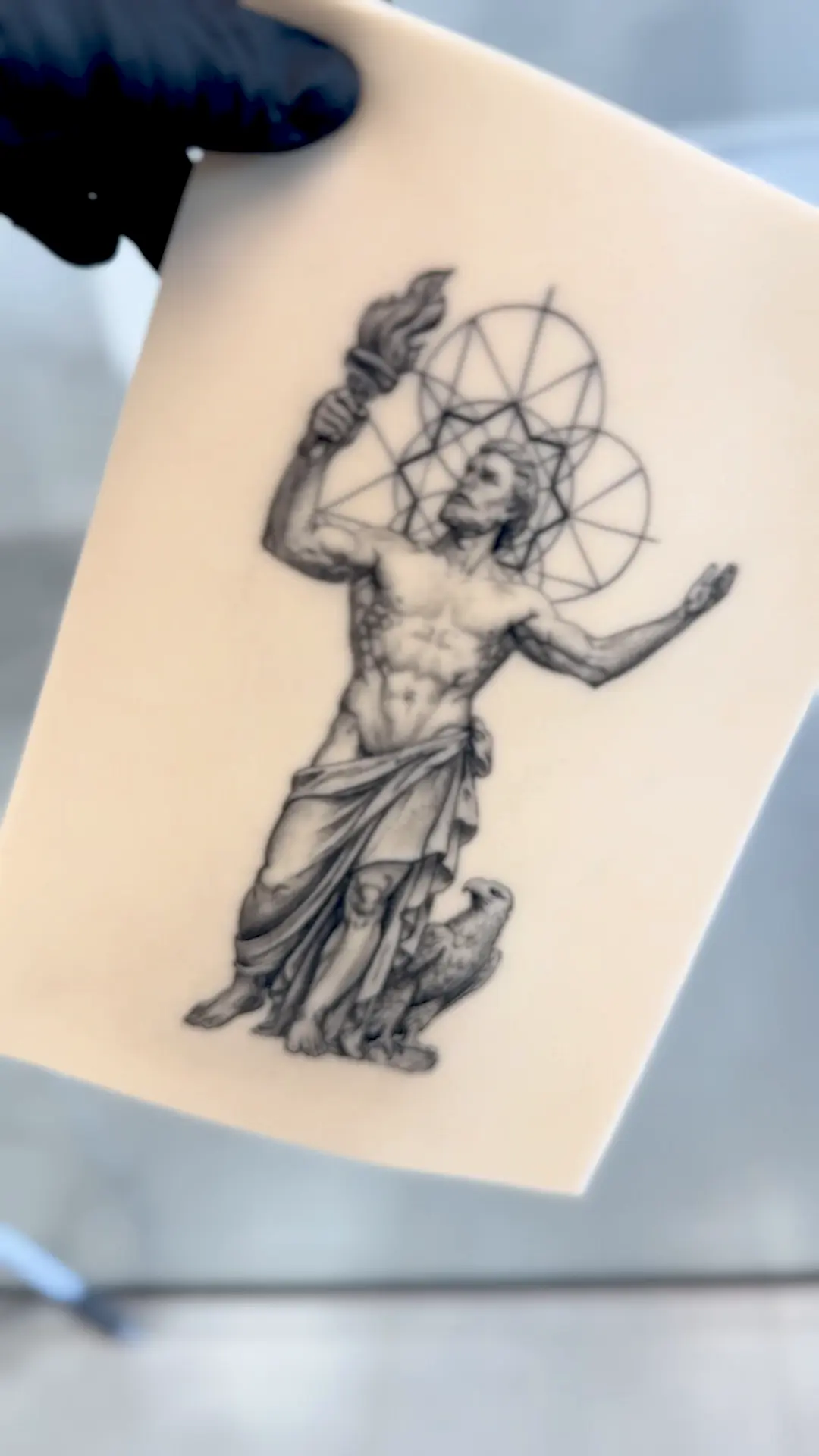 Figura clásica con antorcha y halo geométrico tatuada sobre piel de práctica, sostenida con guante negro