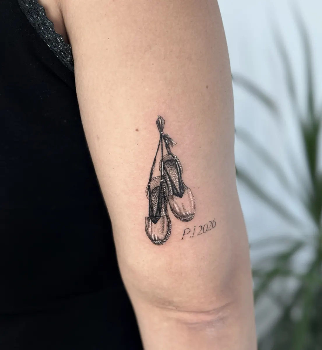 Zapatillas de ballet colgando de sus cintas, tatuadas en microrrealismo en el tríceps, con la fecha en serif fino