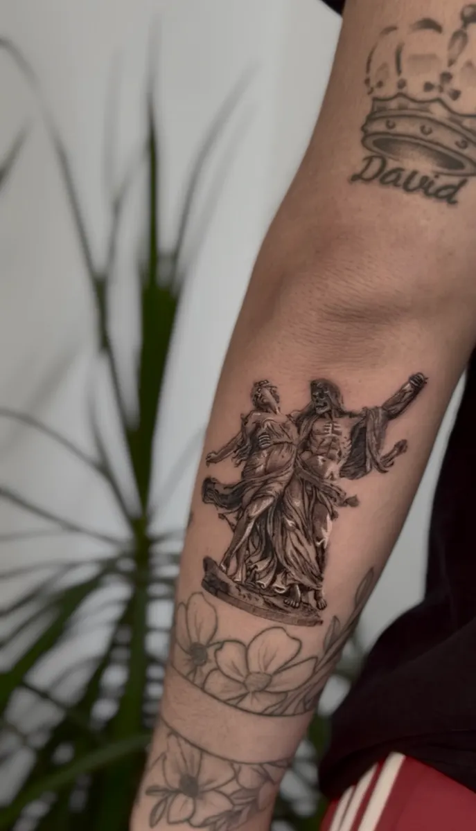 Grupo escultórico neoclásico tatuado en realismo en el antebrazo, sobre un brazalete de flores en línea fina