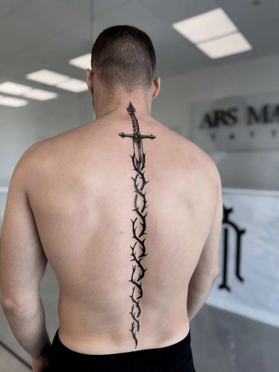 Espada larga envuelta en una rama de espinas, tatuada en blackwork a lo largo de la columna