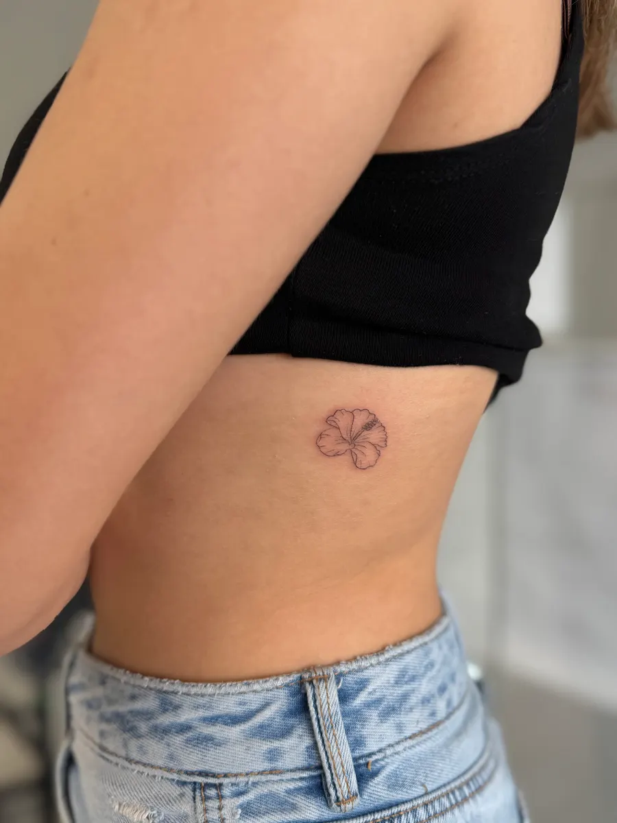 Flor pequeña de cinco pétalos tatuada en línea fina sobre las costillas