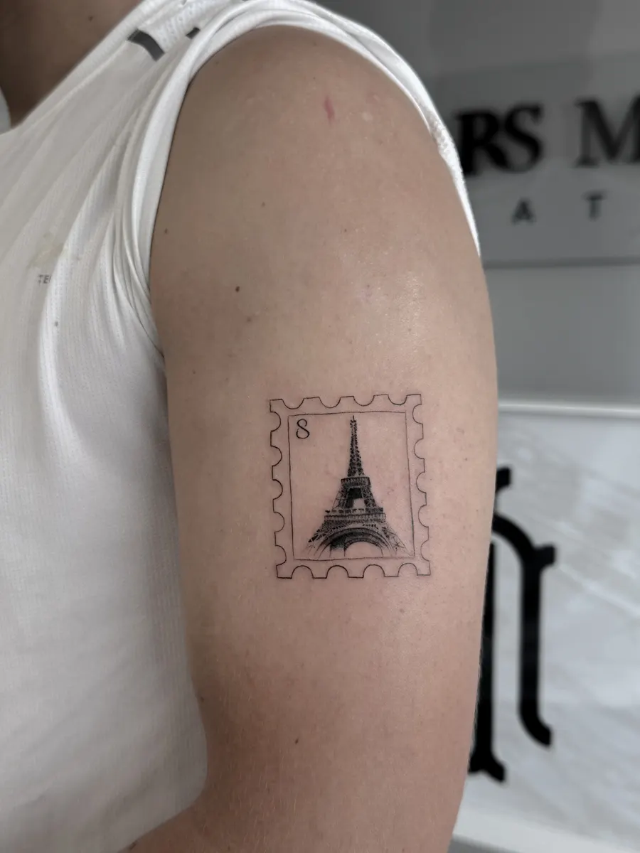 Sello de correos con la torre Eiffel tatuado en línea fina y puntillismo en el hombro