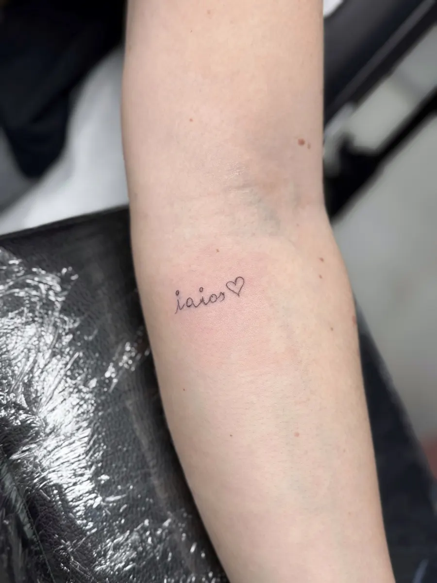 Palabra "iaios" con un corazón, tatuada en cursiva minúscula en el antebrazo